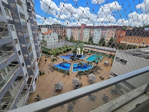 A view of a residential building with multiple balconies, a swimming pool area with sun loungers and umbrellas, and a clear blue sky. - Aptos Rio Quente - ótima área de lazer (Rio Quente)