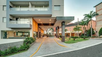 Una entrada de hotel moderna con un corredor cubierto, varios balcones y una zona ajardinada con palmeras.