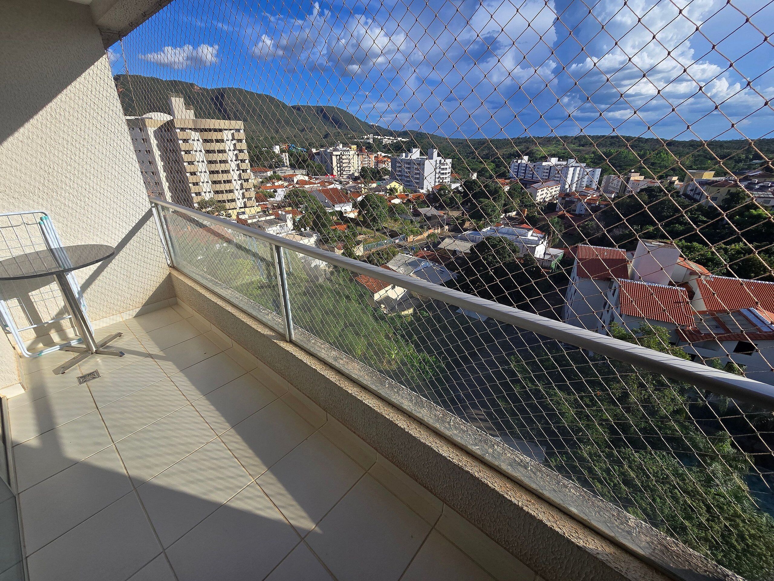 Un balcón con una silla de metal, suelo de baldosas y vistas a edificios y colinas a través de una cerca de alambre.