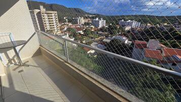 Un balcón con una silla de metal, suelo de baldosas y vistas a edificios y colinas a través de una cerca de alambre.