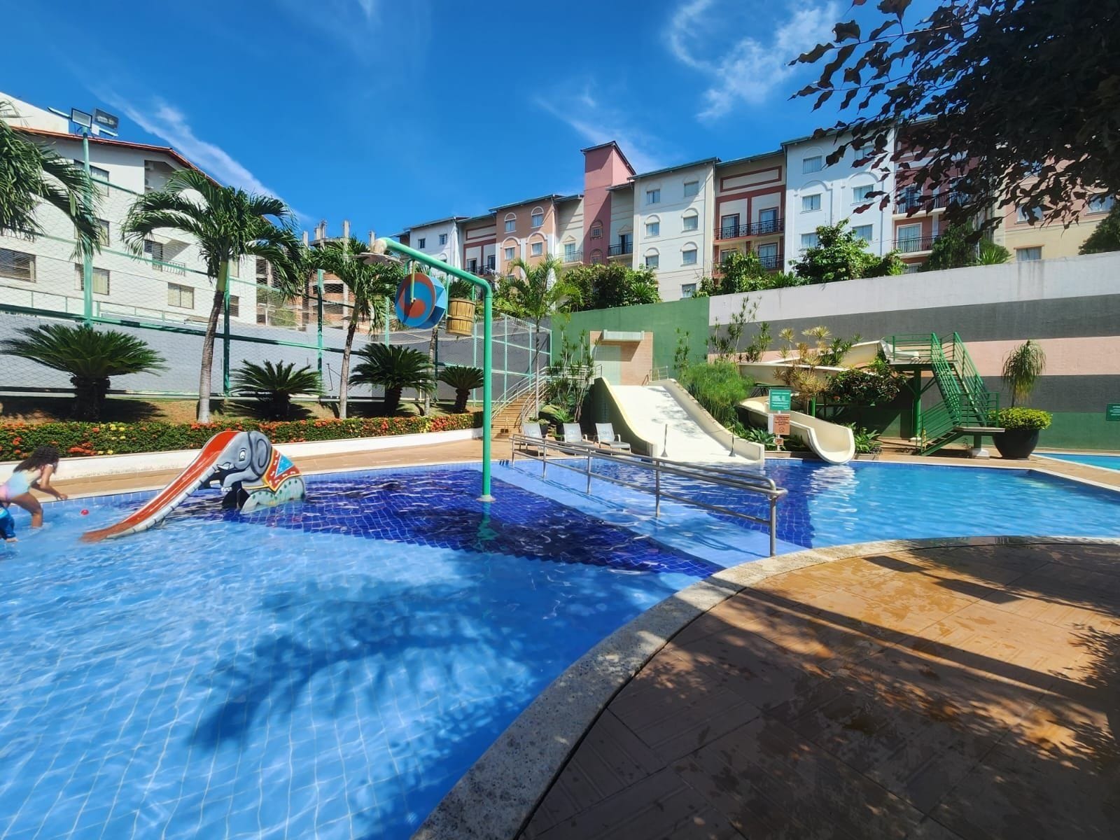 A swimming pool with slides and a children's play area, surrounded by buildings and greenery.