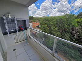 Un balcón con baranda de metal, un trapeador apoyado contra la pared y una vista de un área verde con árboles y edificios al fondo.