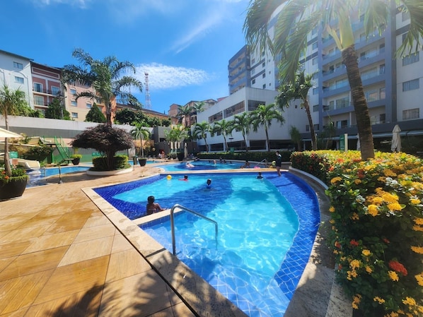 Zona de piscinas con varias piscinas, palmeras y un edificio al fondo.