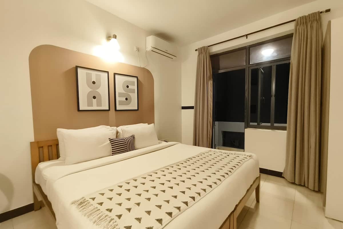 A hotel room with a bed, a headboard, a wall-mounted air conditioner, a window with curtains, and two framed pictures on the wall.