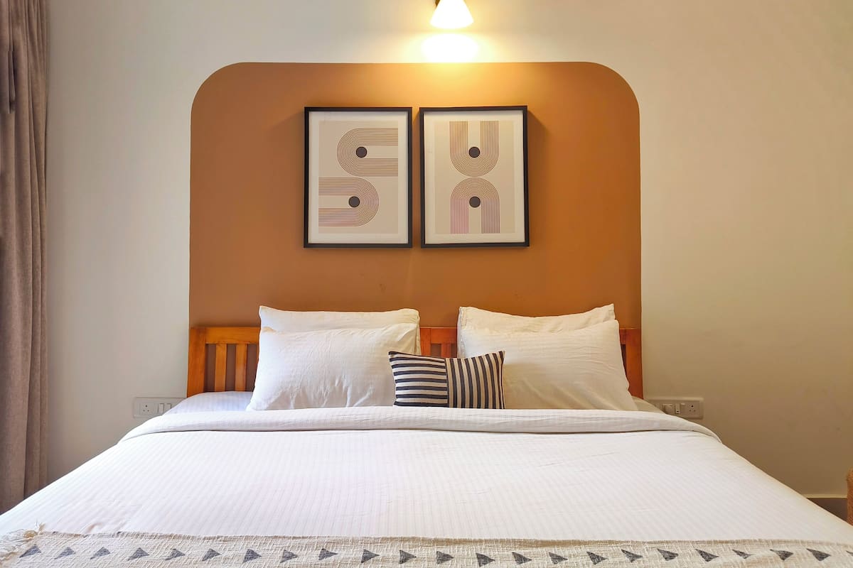 A neatly made bed with white linens and two pillows, a headboard, and two framed artworks on the wall above.
