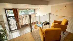 Une pièce avec un fauteuil jaune, une petite table, une vue sur l’extérieur par une fenêtre et un espace clos recouvert d’un filet.