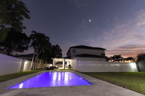 A swimming pool with a blue-lit edge, a white fence, and a house with a satellite dish. - Miami Getaway Heated Pool House & Pool Table Villa (Miami)