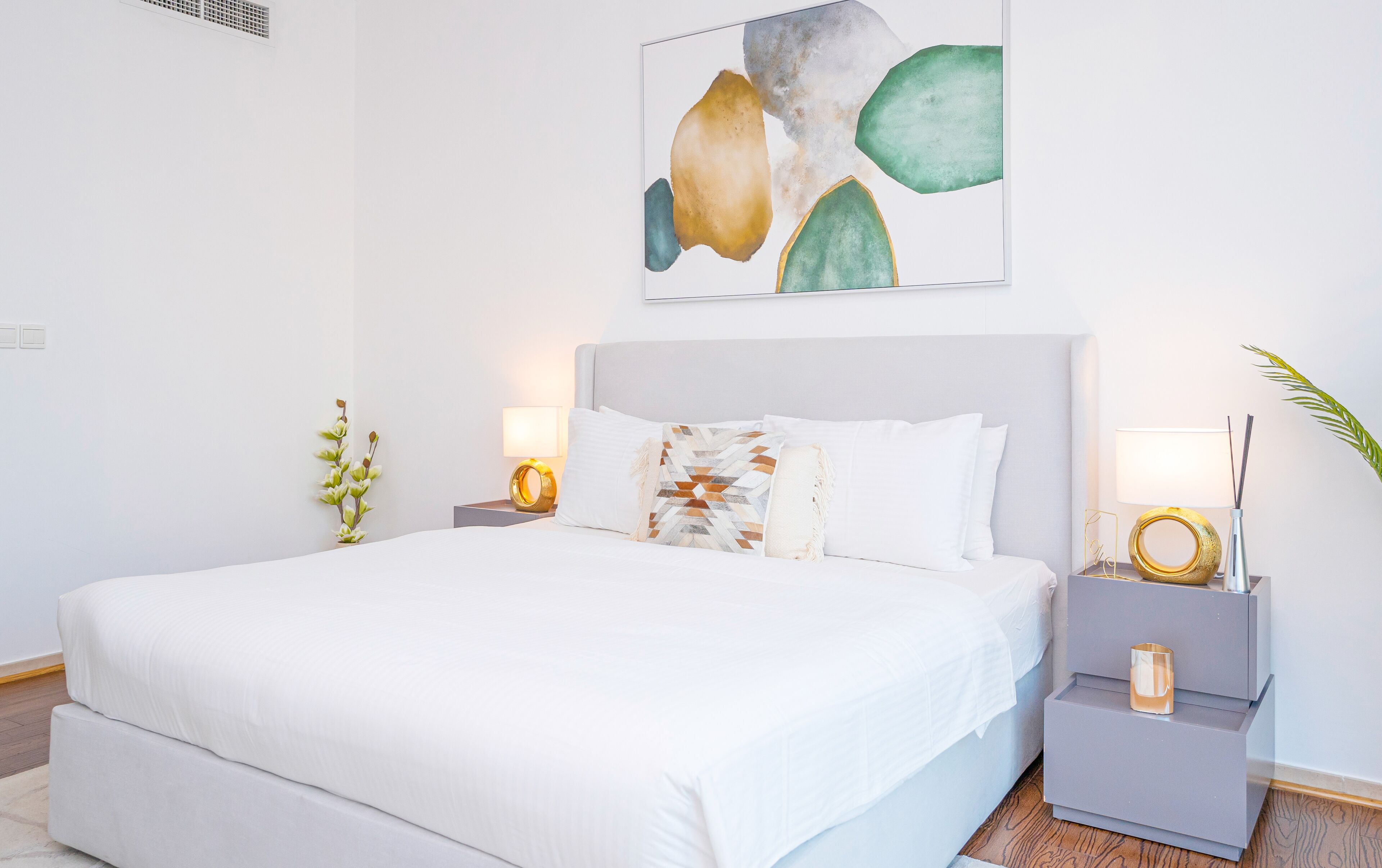 A modern bedroom with a large bed, white bedding, two bedside tables with lamps, and a wall-mounted abstract painting.