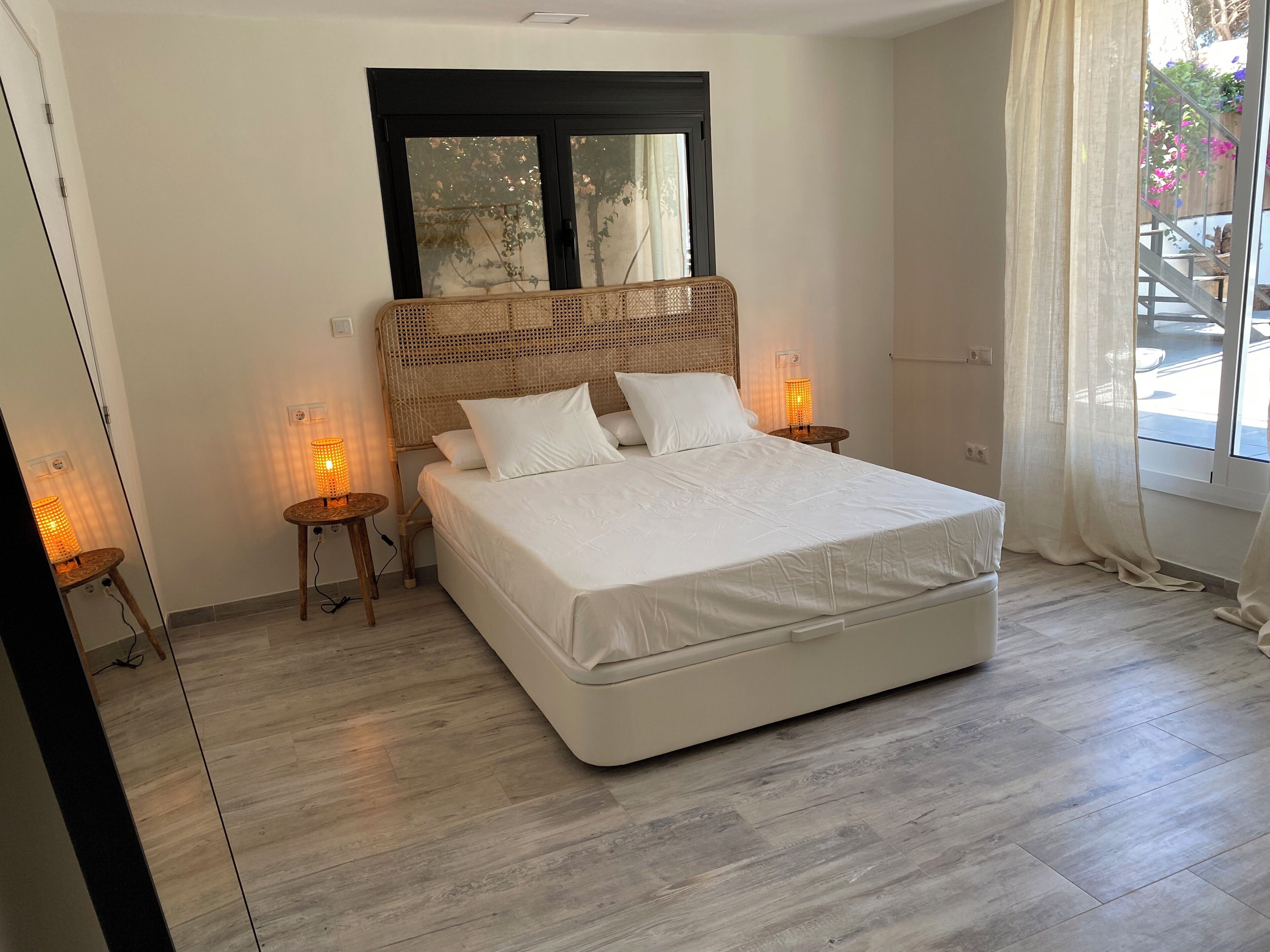 A bedroom with a large bed, two bedside tables with lamps, a window with a black frame, and a view of the outside.