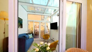 A modern living room with a blue sofa, a yellow chair, and a wicker chair. There is a glass door leading to a balcony with a wicker chair and a TV mounted on the wall. - Live the Magic of Taormina Where History, Beauty, and Sicilian Soul Meet (Taormina)