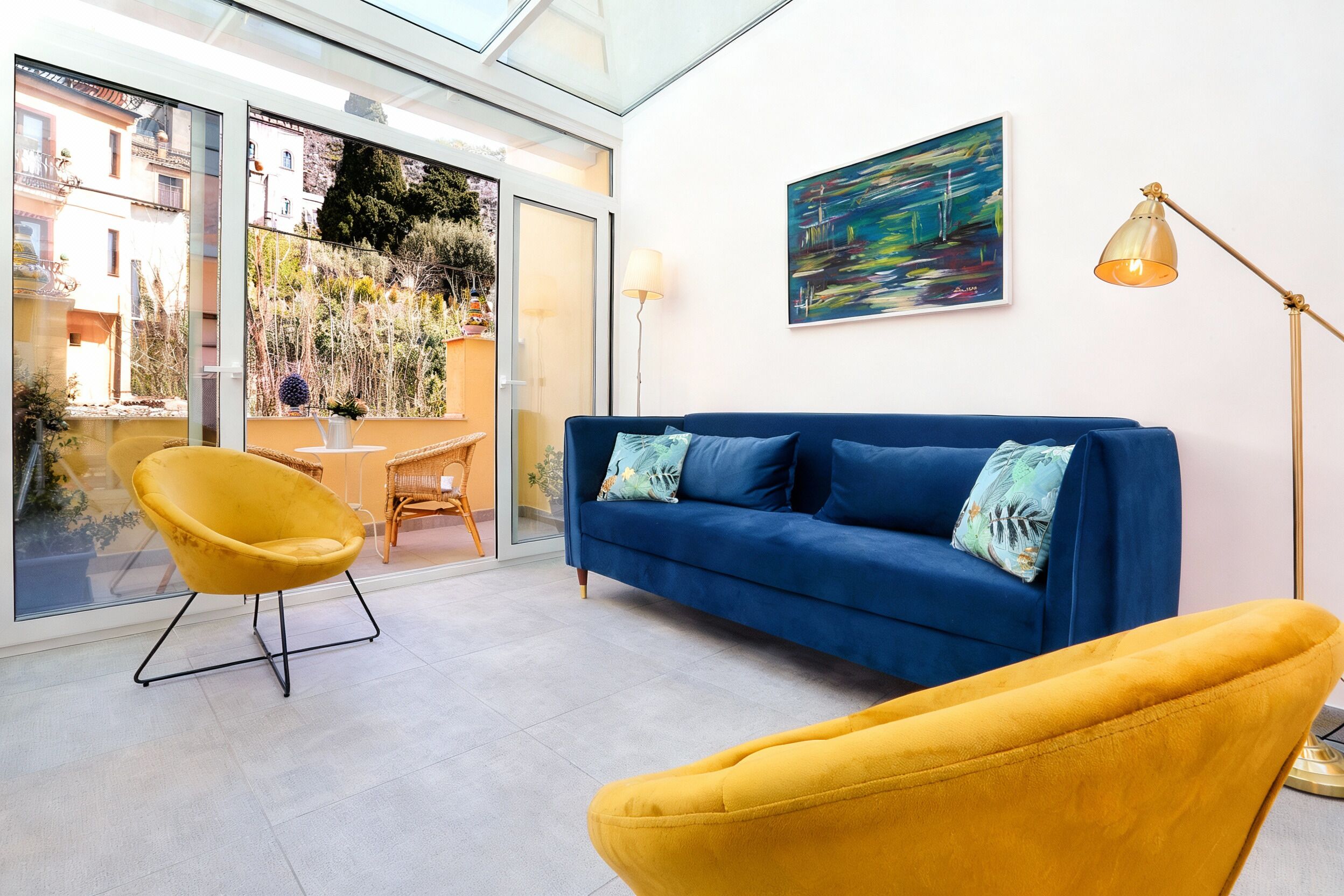 A modern living room with a blue sofa, yellow armchair, and a glass door leading to an outdoor area with plants and a view of buildings.