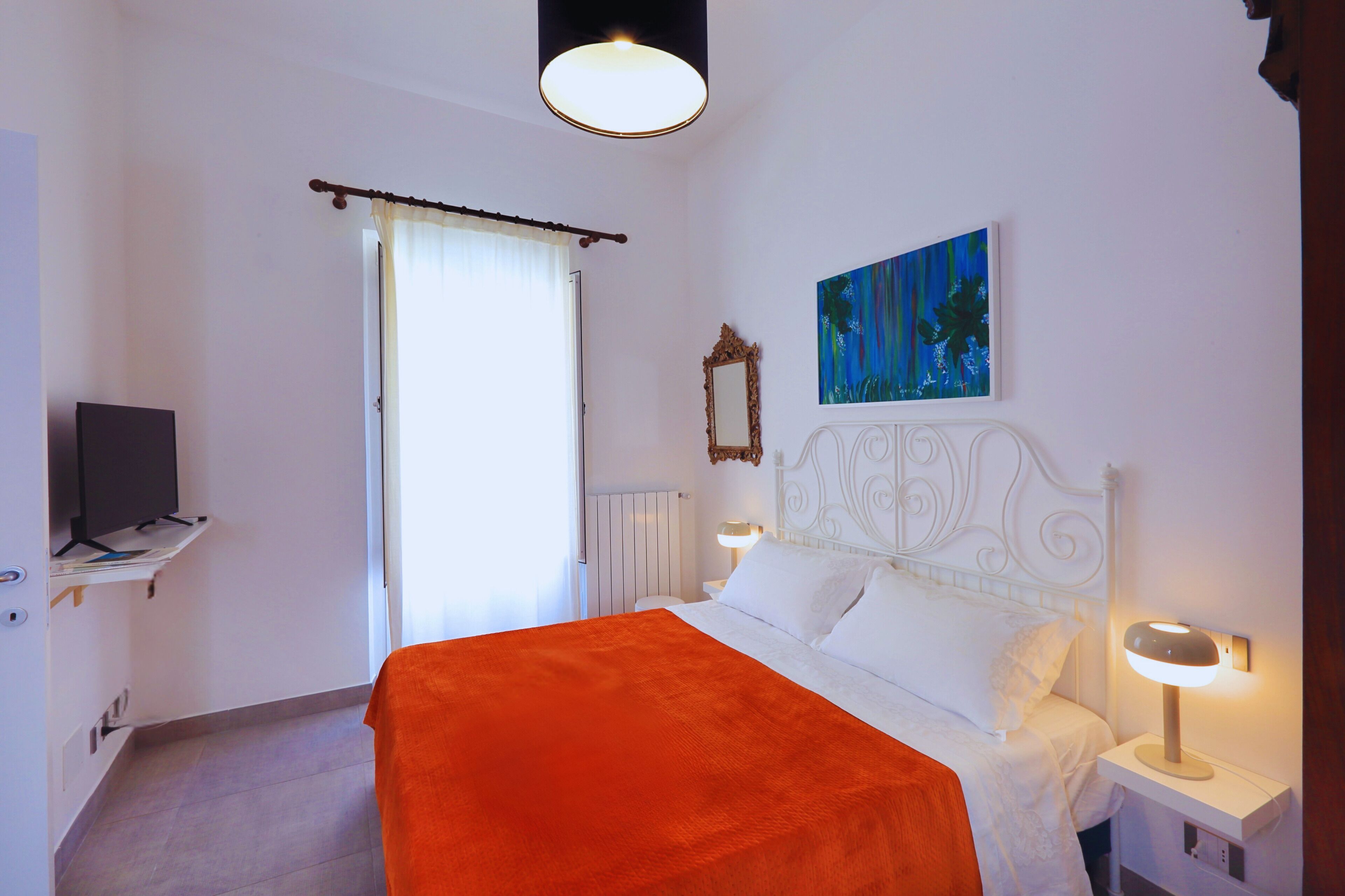 A hotel room with a bed, a television on a shelf, a painting on the wall, and a window with curtains.