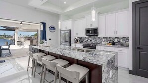 A modern kitchen with white cabinets, a marble island with bar stools, a stainless steel refrigerator, and a microwave.