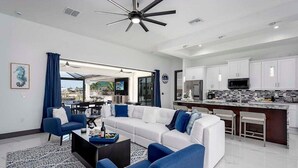 A modern living room with a white sectional sofa, blue armchairs, and a coffee table. A kitchen with white cabinets and a bar area is visible in the background.