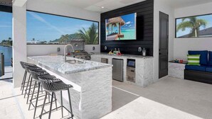 A modern outdoor kitchen with a large island, bar stools, a TV, and a view of the water.