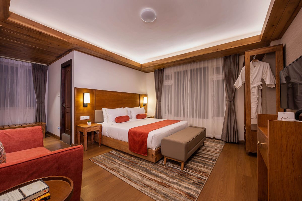A hotel room with a wooden ceiling, a large bed with a red blanket, a wooden bench, a red armchair, a small table with a lamp, and a mirror.