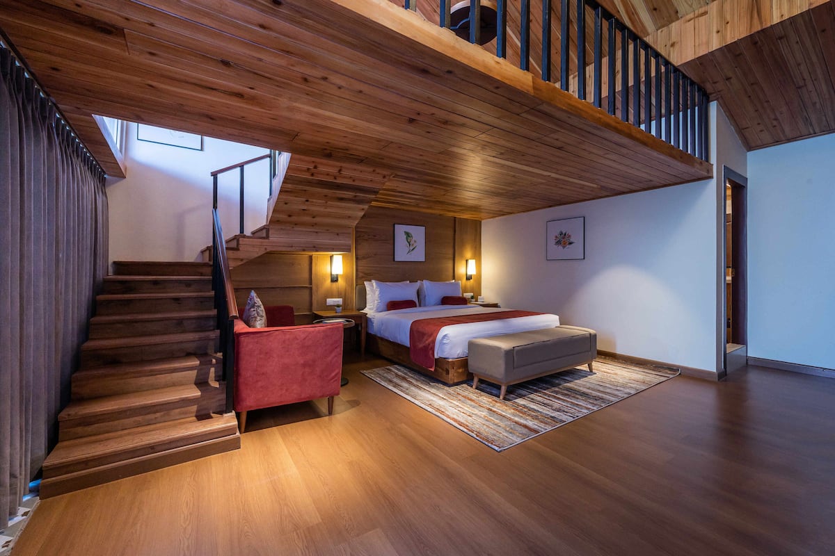 A spacious bedroom with a wooden staircase, a bed with red bedding, a red armchair, and a bench.