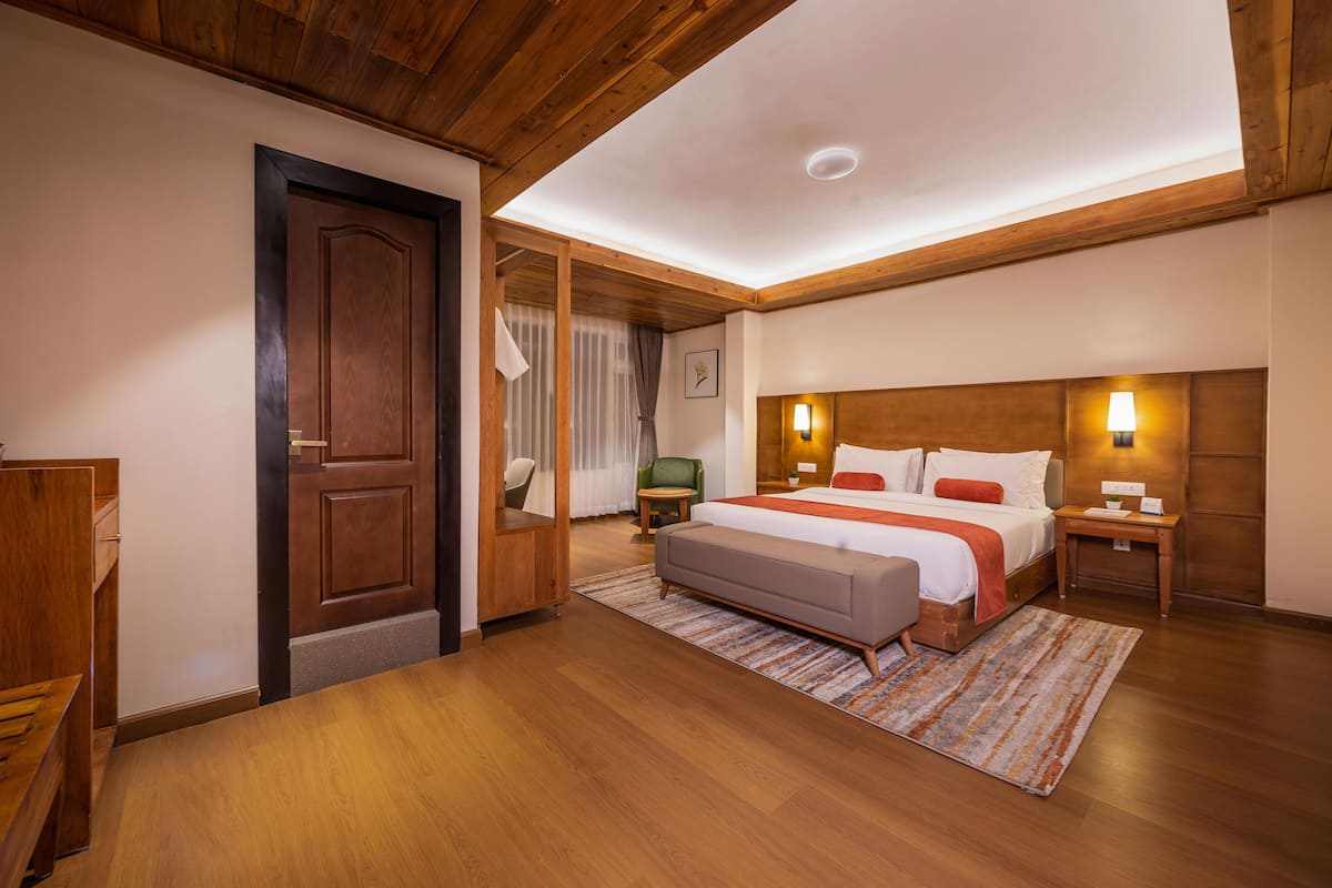 A hotel room with a wooden ceiling, a large bed with a headboard, a bedside table, a nightstand, a chair, a small table, and a door.