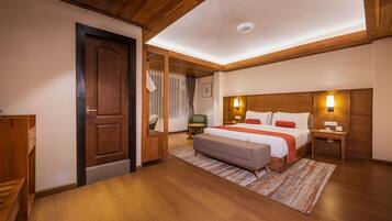 A hotel room with a wooden ceiling, a large bed with a headboard, a bedside table, a nightstand, a chair, a small table, and a door.