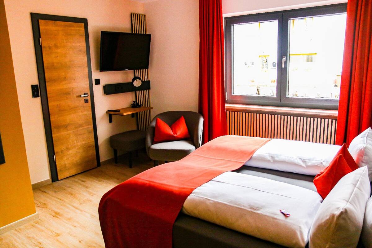 A hotel room with a bed, a TV mounted on the wall, a chair with a red cushion, and a window with red curtains.