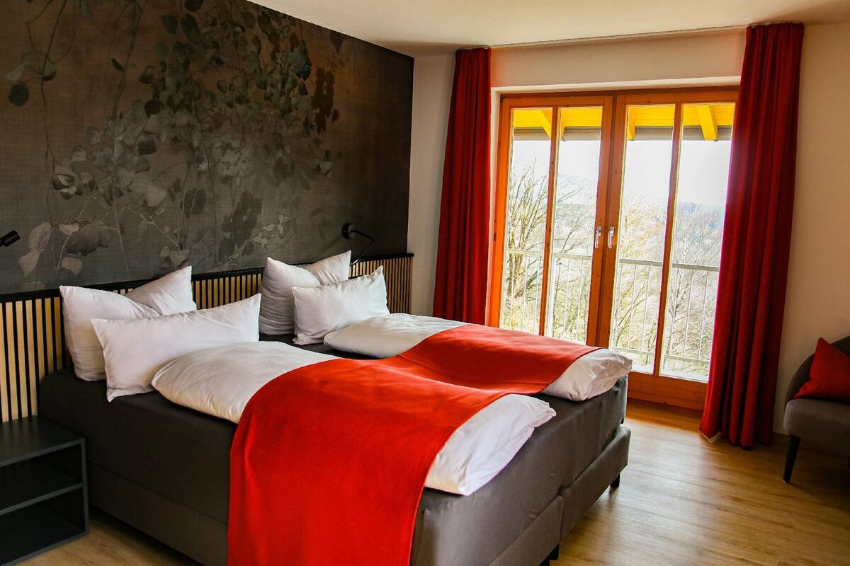 A modern bedroom with a large bed, a view of trees through a sliding glass door, and a red curtain.