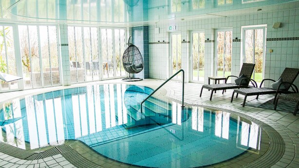 An indoor swimming pool with clear glass walls, lounge chairs, and a hanging chair. - DAS PALMBERGER (Spiegelau)