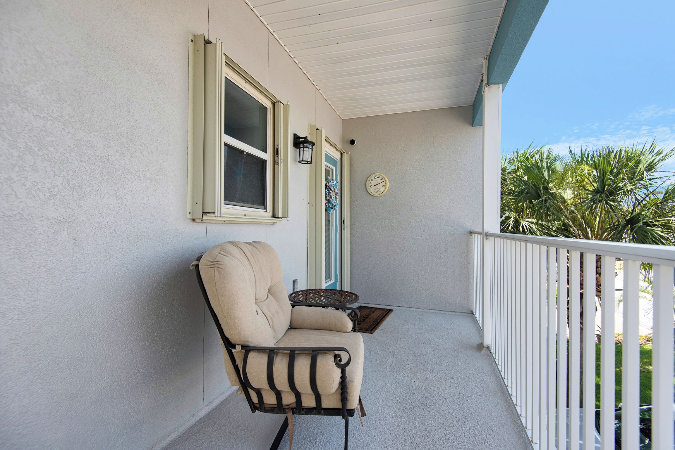 A covered porch with a beige chair, a small table, and a wall-mounted clock.