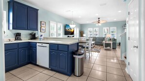 Een moderne keuken met blauwe kastjes, een vaatwasser en een vuilnisbak. Er is een eethoek met een tafel en stoelen, en een woonkamer met een bank en een televisie.
