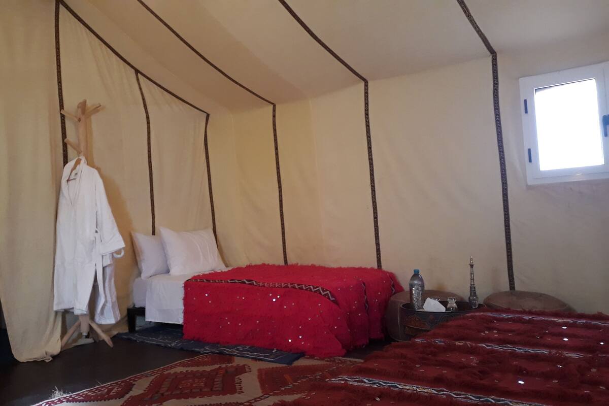 Une pièce avec un lit, une couverture rouge, une robe blanche suspendue et une petite table sur laquelle se trouve une bouteille.