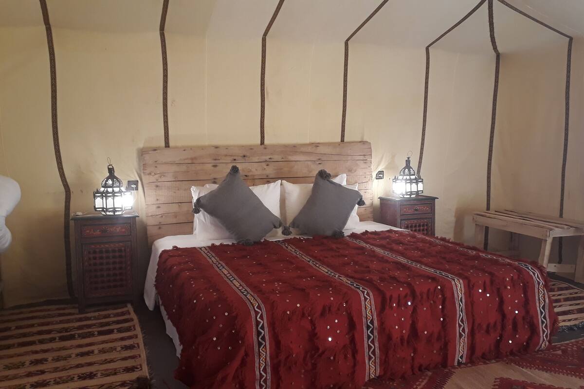 Une chambre avec une tête de lit en bois, un lit recouvert d’une couverture rouge, deux tables de chevet ornées de lanternes et un tapis à motifs.
