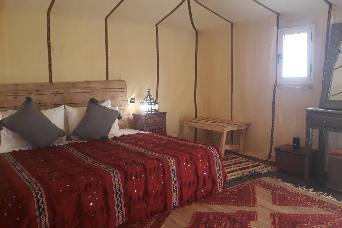 Une chambre à coucher avec une tête de lit en bois, un lit avec une literie rouge, une table de chevet avec une lanterne, une petite table, un miroir et une cheminée.