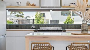 A modern kitchen with a central island, two wicker chairs, a stainless steel range hood, and a view of the neighborhood.