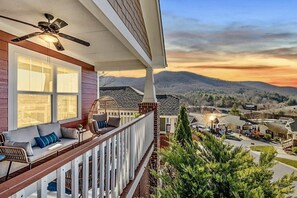 Een balkon met een plafondventilator, een bank met kussens, en uitzicht op een woongebied met huizen en bergen op de achtergrond.