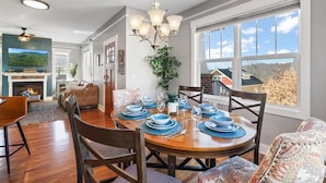 A dining area with a wooden table set for six, surrounded by chairs. A chandelier hangs above, and the room has large windows offering a view of the outdoors. - Mountain Views, Game Room, Hot Tub –Close to Downtown (Asheville)