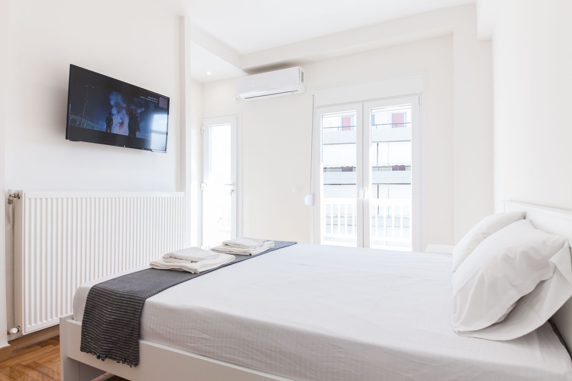 A bedroom with a bed, a TV mounted on the wall, a radiator, and a balcony visible through the door.