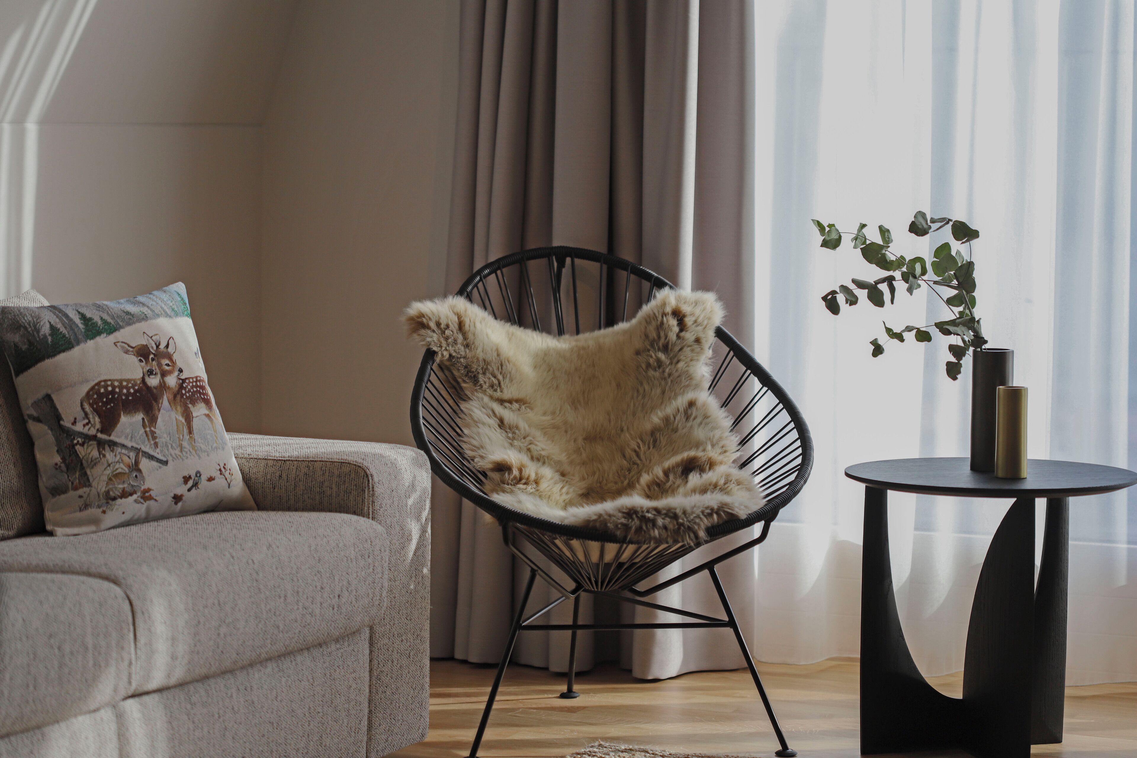 Una sala moderna con un sofá gris, una silla de estructura metálica negra con una manta de piel, una mesita redonda con una planta y cortinas traslúcidas.
