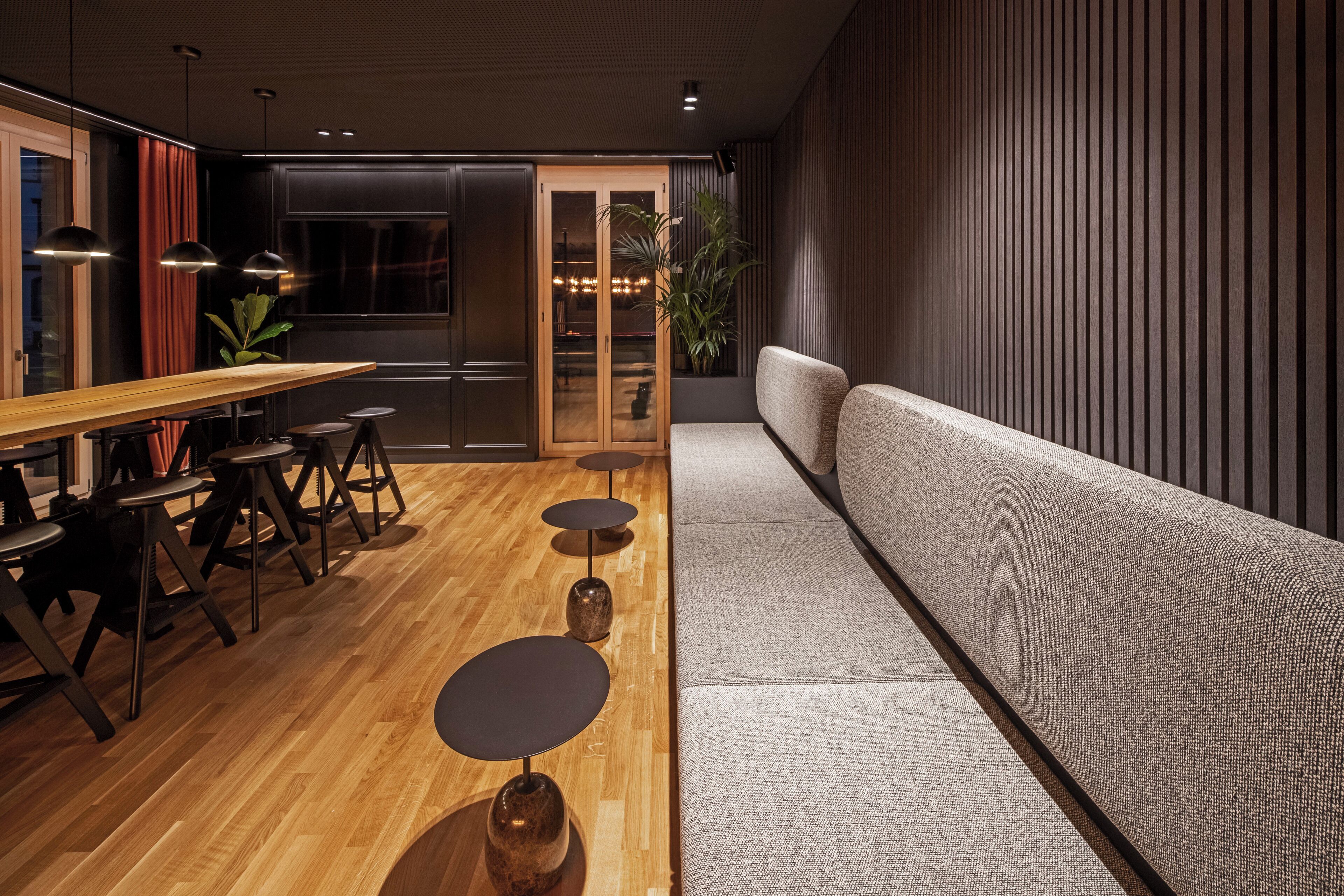 A modern interior with a wooden dining table, black stools, a grey sofa, and a large window with curtains.