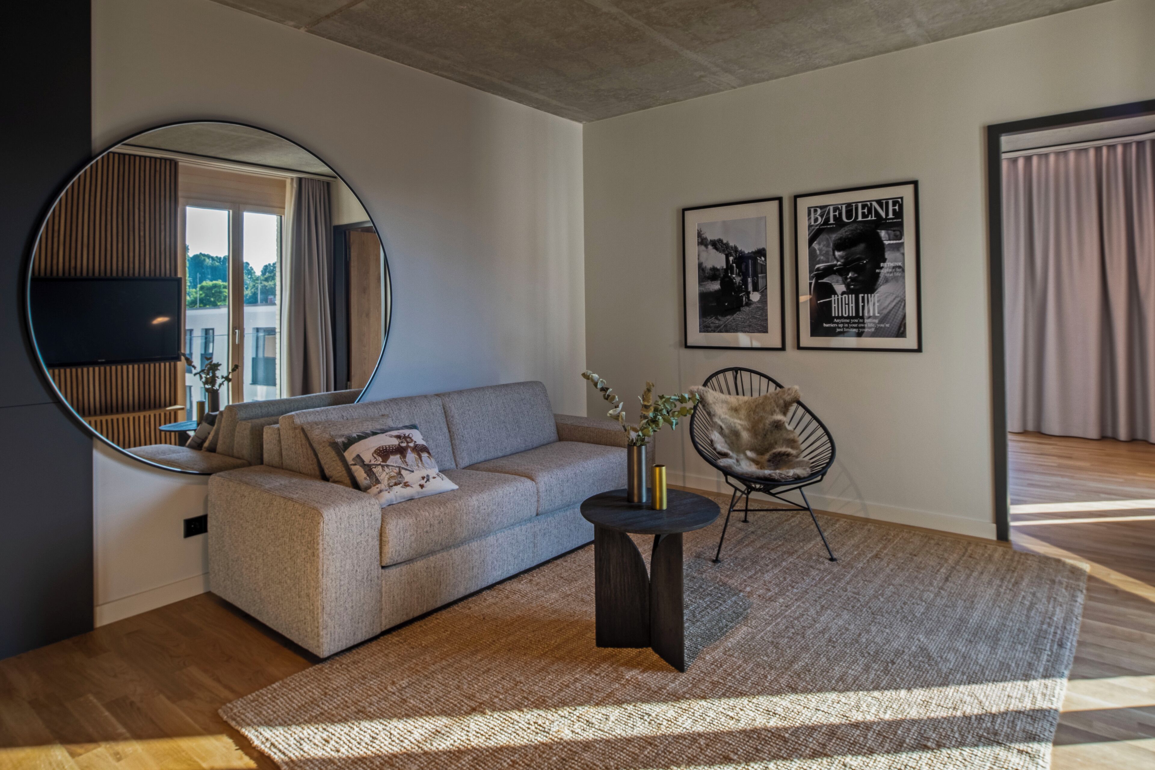 A modern living room with a sofa, a round mirror, a coffee table, and a chair. There are framed pictures on the wall and a window with curtains.