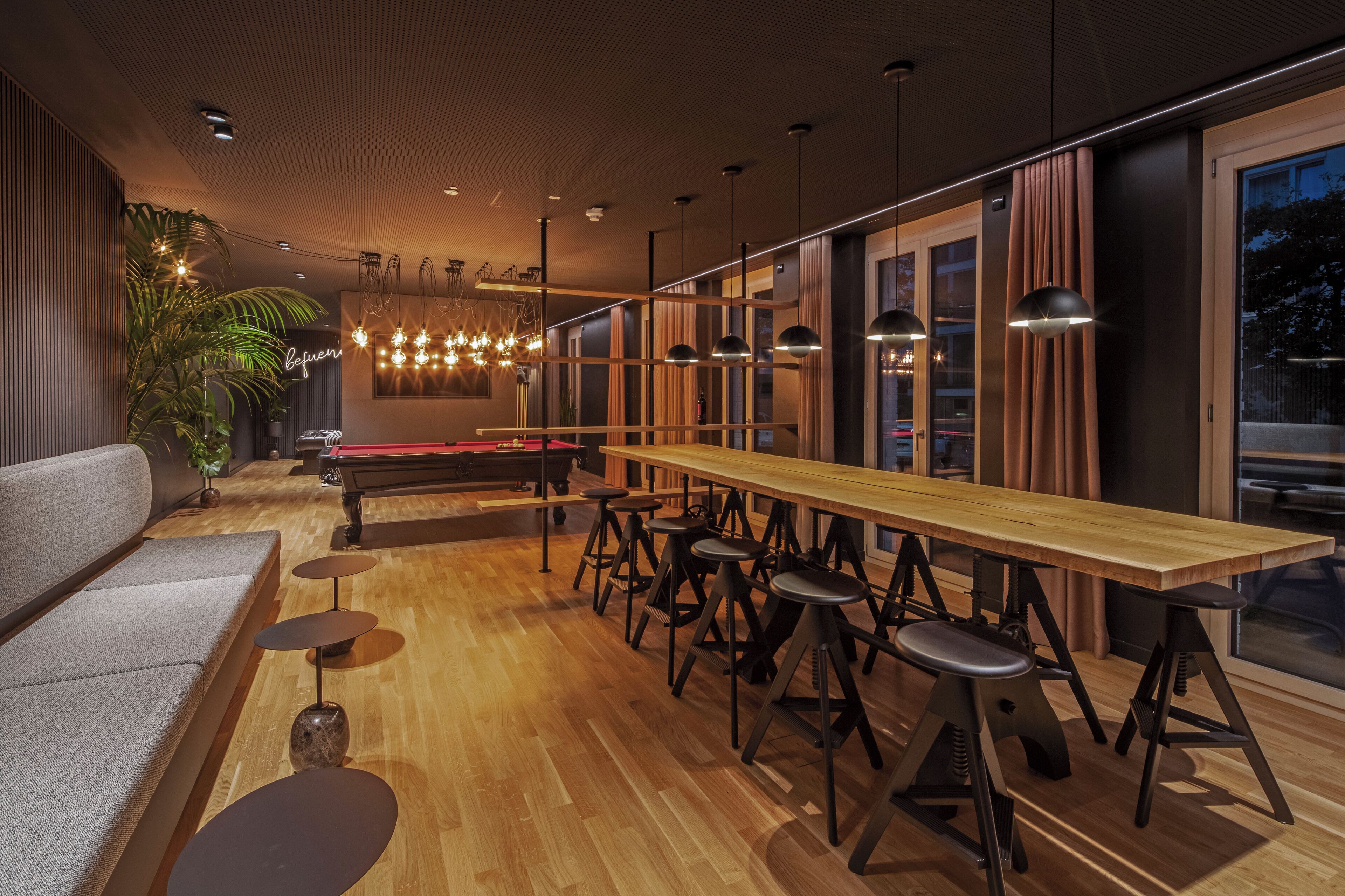 A modern bar area with a long wooden table, bar stools, and a pool table.