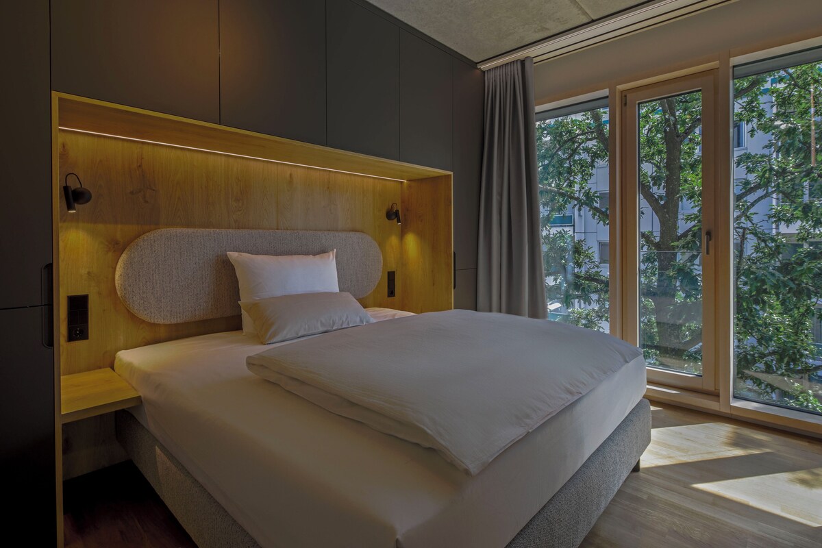 A modern bedroom with a large bed, wooden headboard, and a view of trees through large windows.