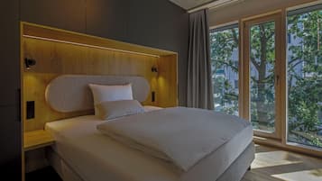 A modern bedroom with a large bed, wooden headboard, and a view of trees through large windows.