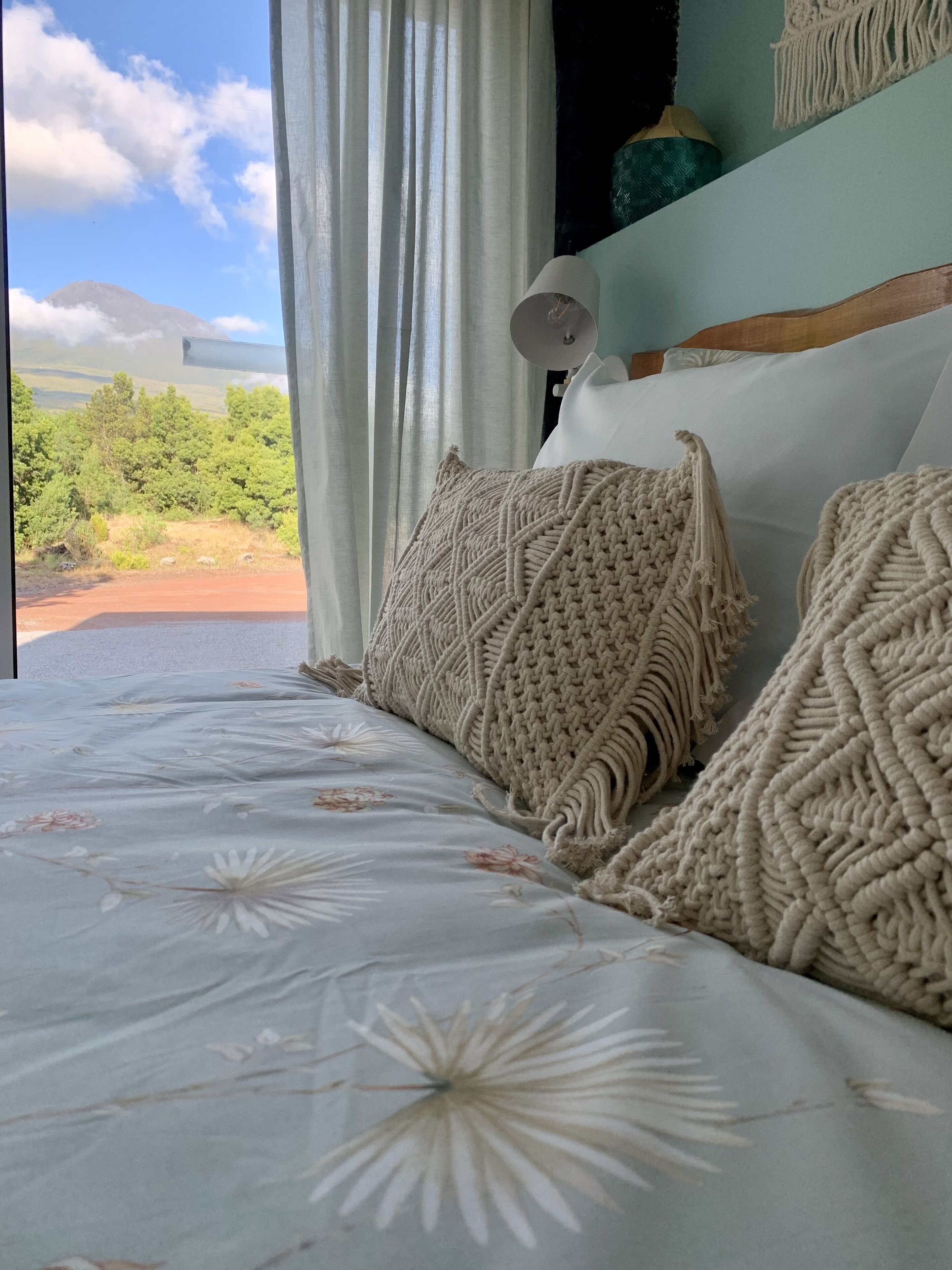 Un lit agrémenté d’oreillers en macramé et d’une couvre-lit à motifs floraux, une décoration murale en macramé, et une vue sur un paysage verdoyant à travers des rideaux transparents.