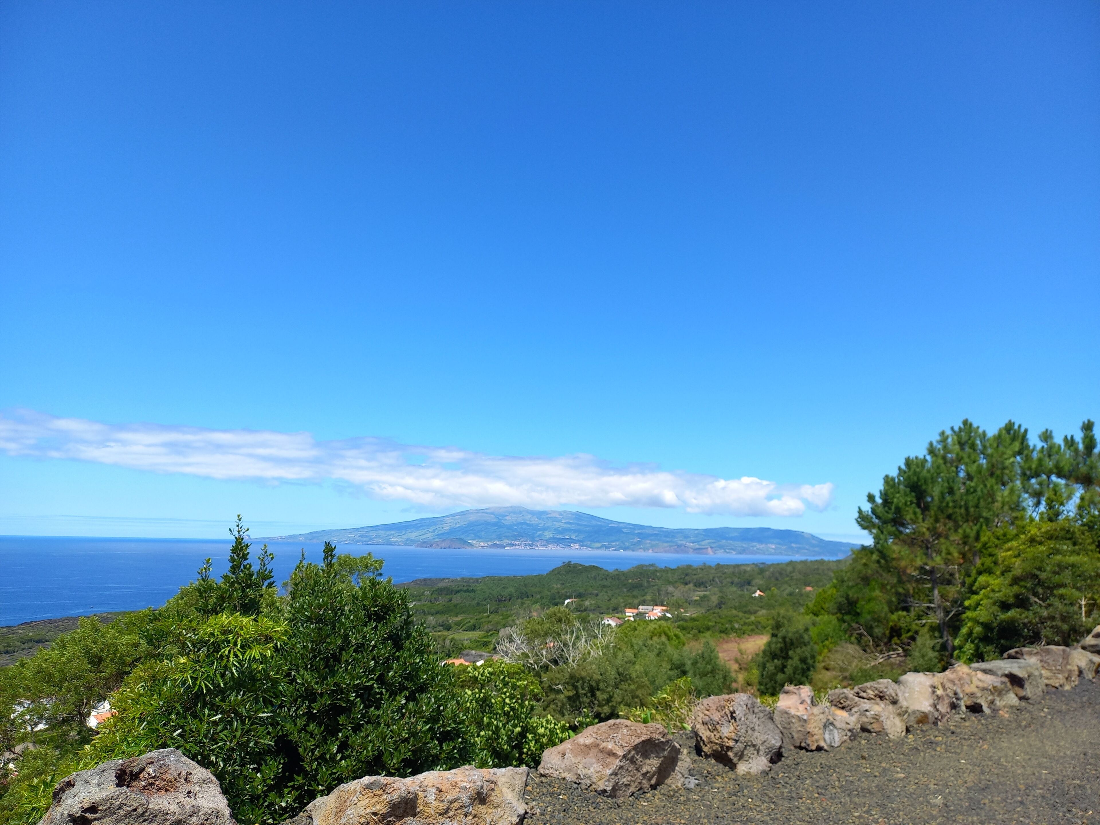 A coastal landscape with rocky terrain, dense greenery, and a distant view of the sea and mountains.
