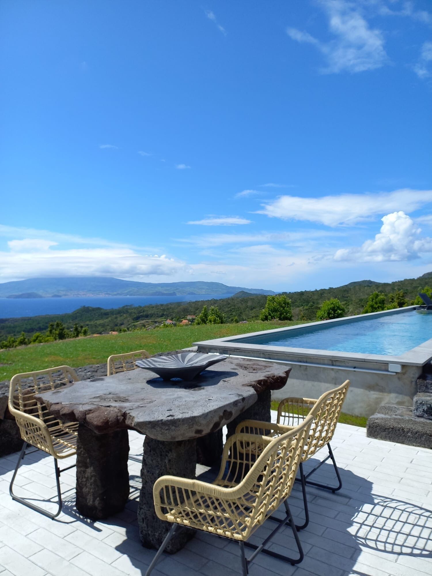 Un espace aménagé au bord de la piscine, avec une table ronde et des chaises en osier, offrant une vue sur un paysage pittoresque composé de collines et d’un ciel dégagé.