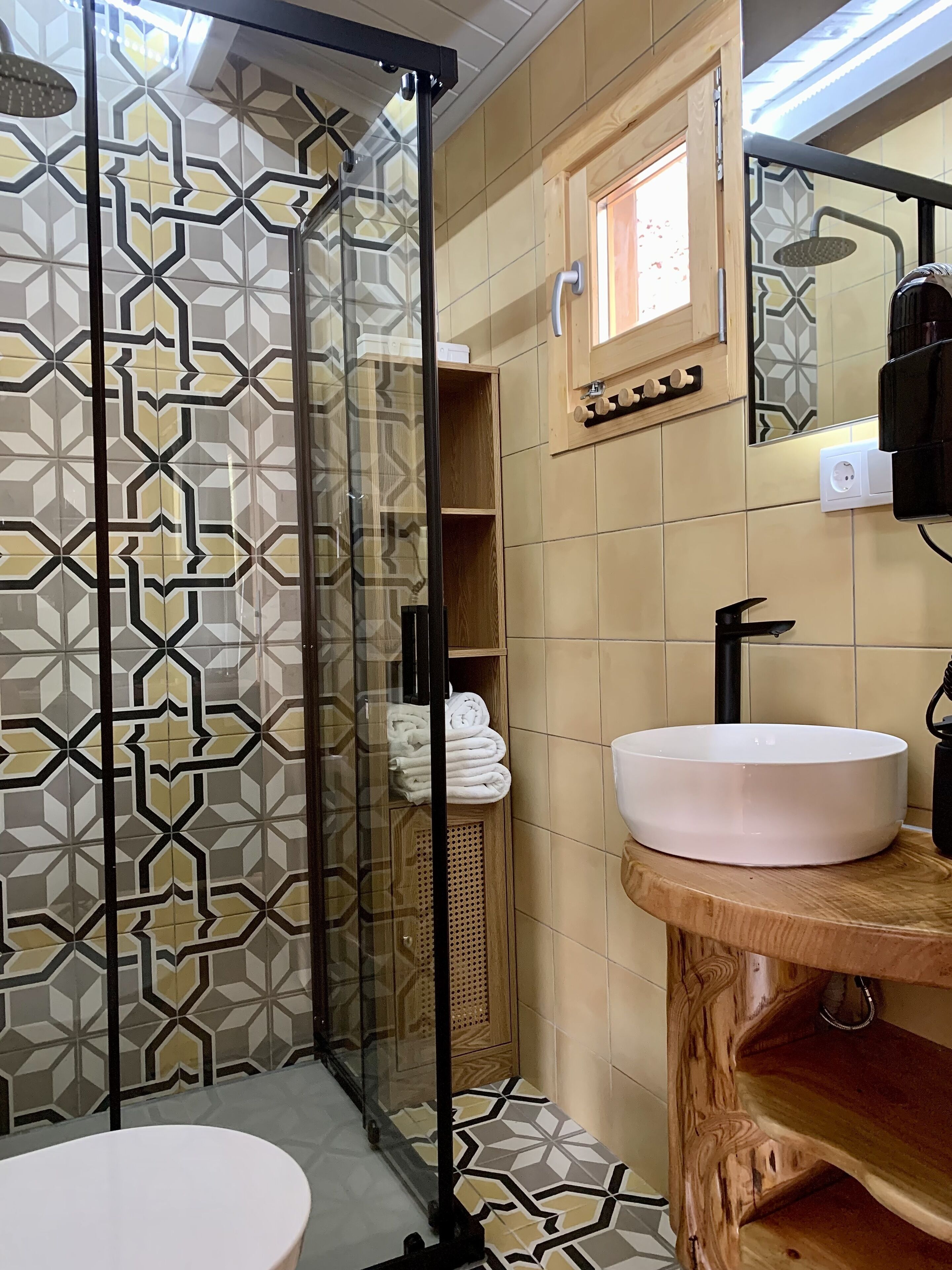 A bathroom with a geometric patterned tile wall, a glass shower enclosure, a wooden vanity with a sink, and a towel rack.