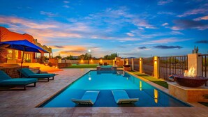 A swimming pool with lounge chairs and a fire feature, surrounded by a fence and a sunset sky.