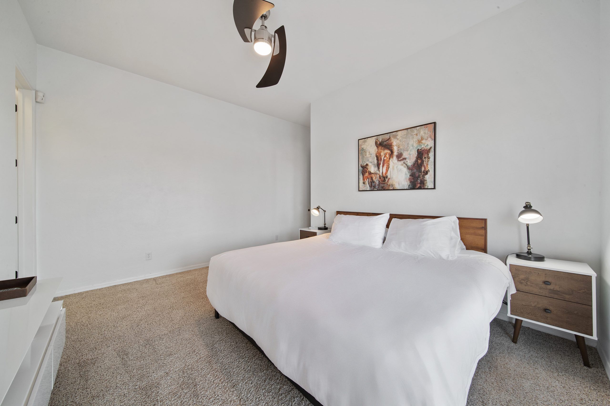 A bedroom with a large bed, two bedside tables with lamps, a wall-mounted artwork, and a ceiling fan.
