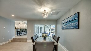 Un comedor moderno con una mesa y sillas de vidrio, una lámpara en el techo, y vista a la cocina y el comedor al fondo.