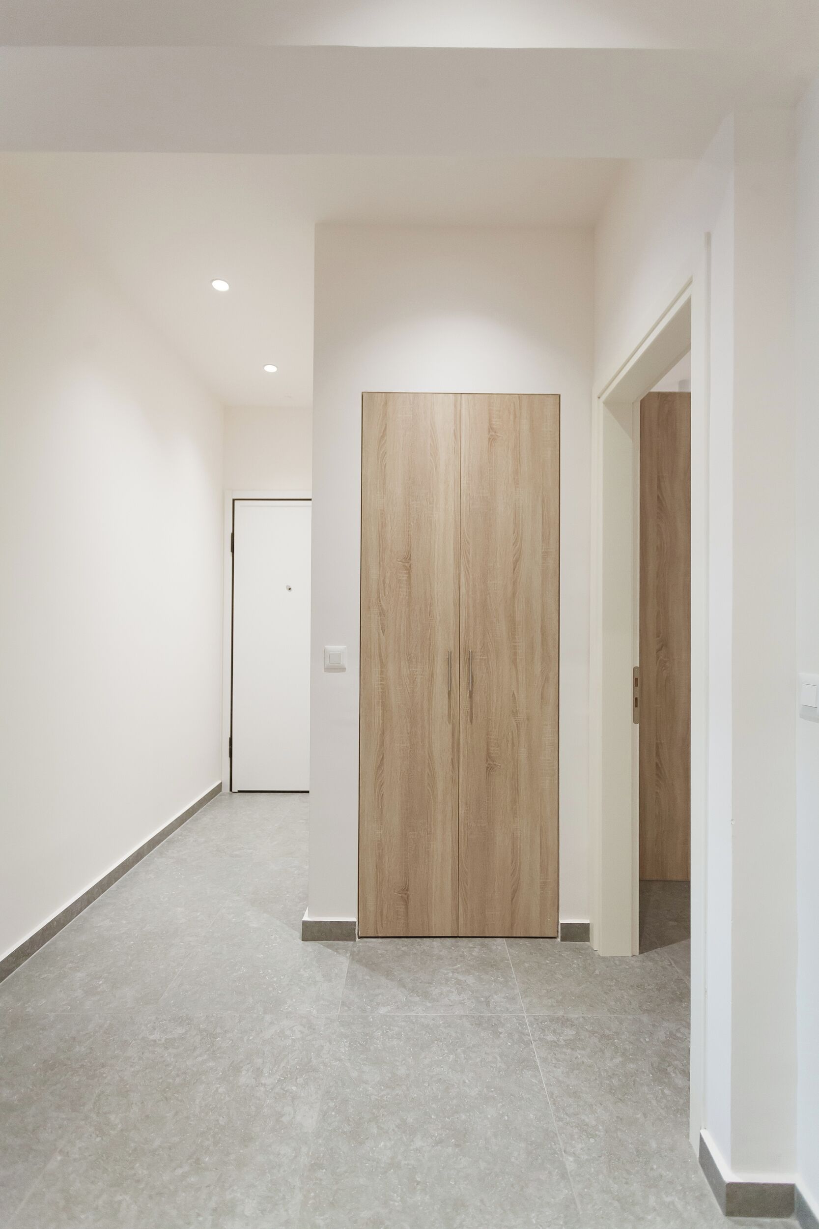 Un pasillo con puerta de madera, paredes blancas y suelo de baldosas.