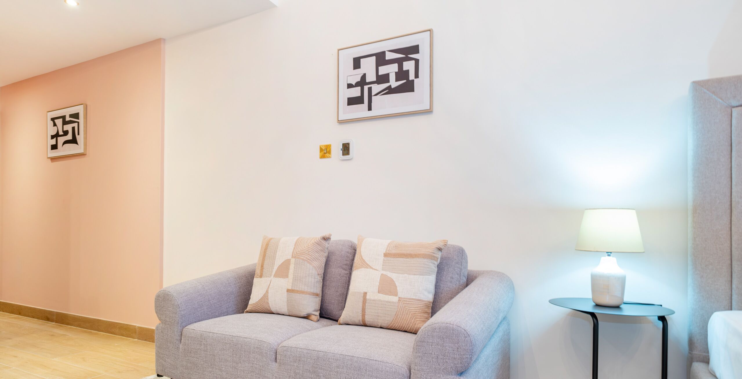 Moderni hotellihuone, jossa on harmaa sohva, koristeellisia tyynyjä, pieni sivupöytä lampun kanssa ja abstraktia seinätaidetta.
